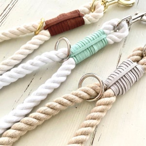 May include: Close-up of four dog leads with thick, twisted rope in cream, white, and tan. Each lead features a leather accent in brown, mint green, and grey, with a metal ring and clasp.