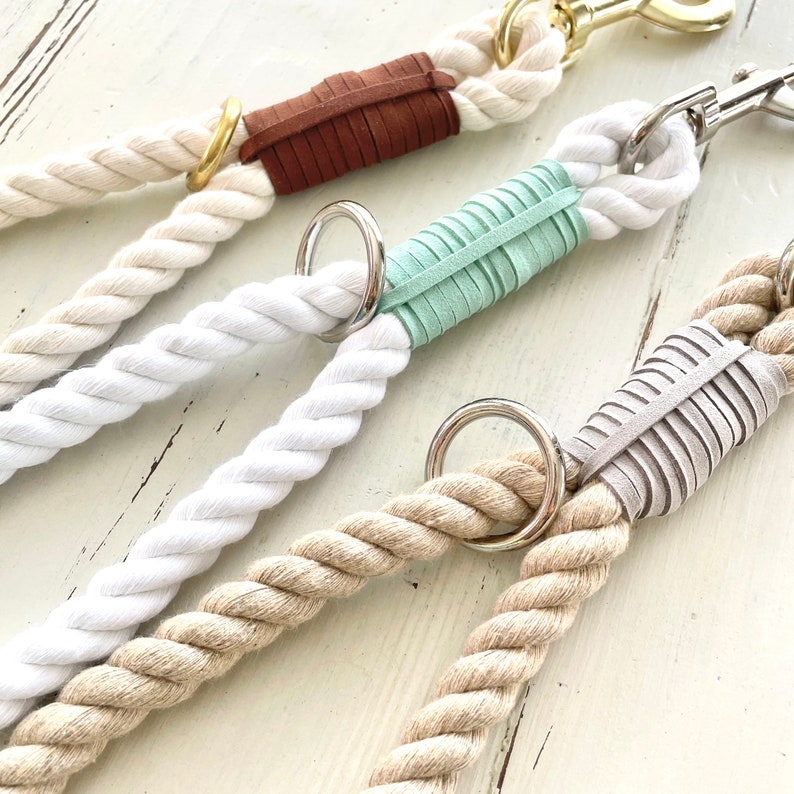 May include: Close-up of four dog leads with thick, twisted rope in cream, white, and tan. Each lead features a leather accent in brown, mint green, and grey, with a metal ring and clasp.