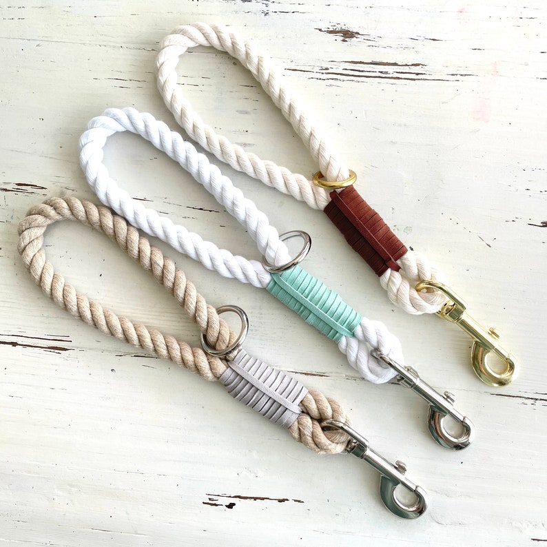 May include: Three dog leads with thick, twisted rope handles. One lead is off-white with a brown leather accent and gold-coloured clasp. The other two are white and tan with silver clasps and mint and grey accents.