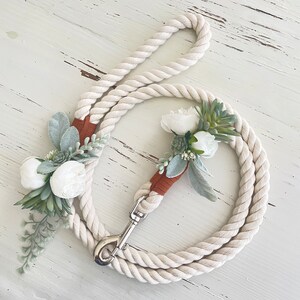 May include: A white cotton rope dog leash with a silver clip and two floral embellishments. The flowers are white and green.