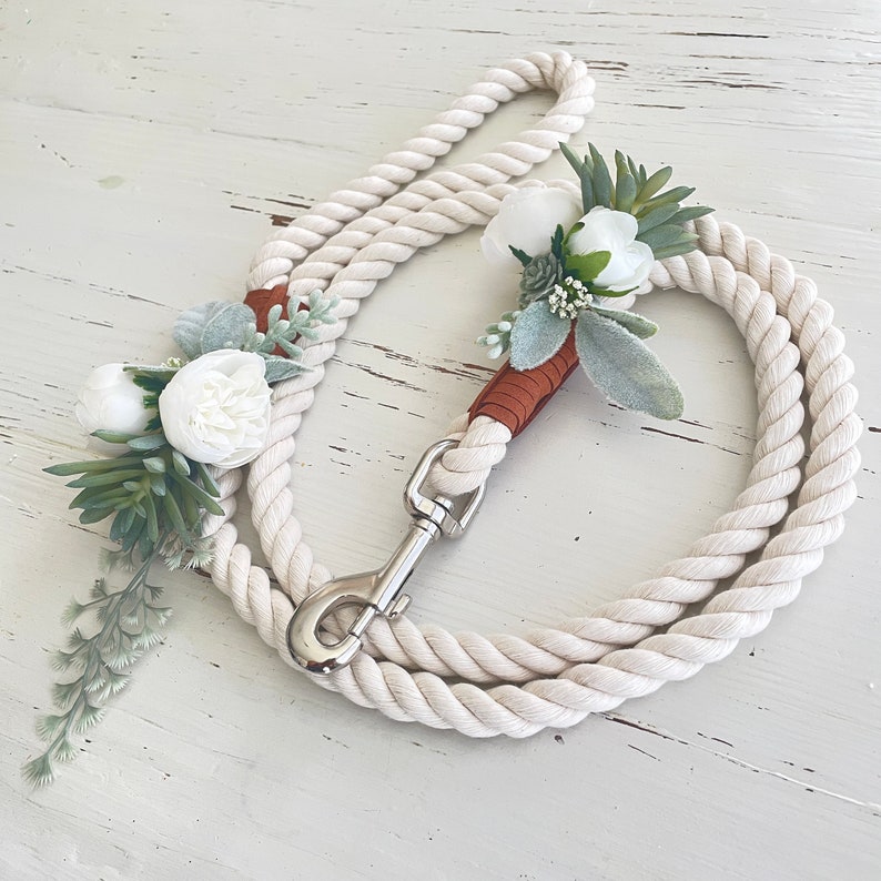 May include: A white rope dog leash with a brown leather handle and a white flower and greenery accent.