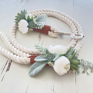 May include: Two white rope leashes with brown leather accents and white flower and greenery embellishments.