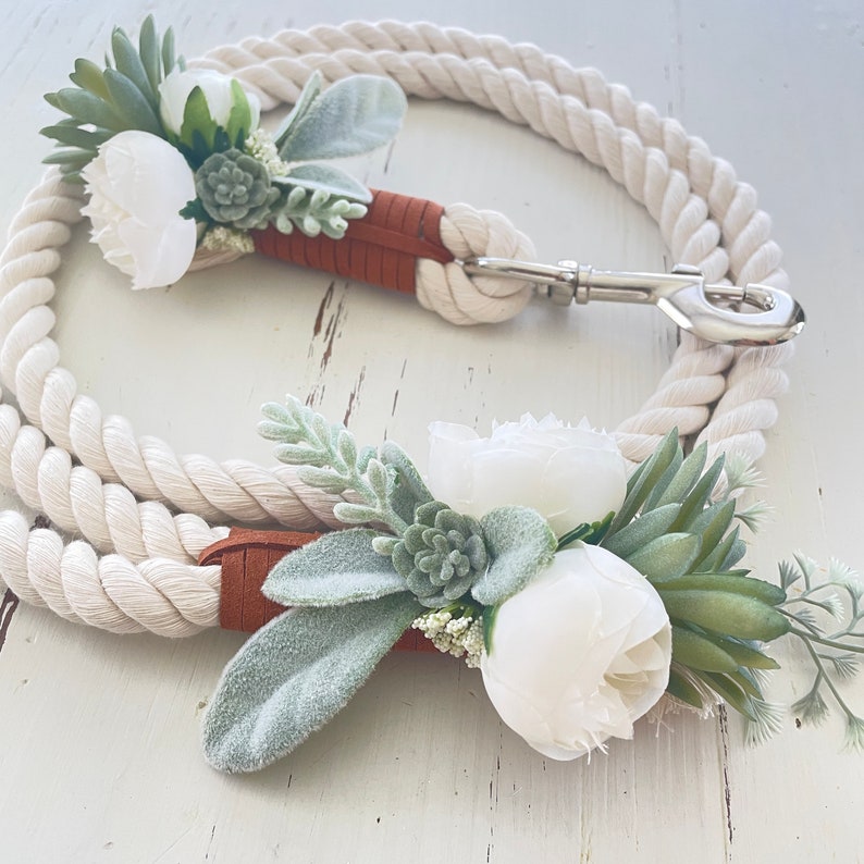 May include: White cotton rope dog leash with a brown leather handle and a silver clip. The leash is decorated with two white flower arrangements with green succulents and leaves.