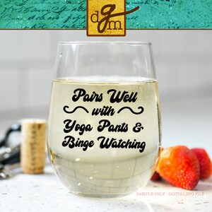以下が含まれることがあります： 白いワインが入った透明なガラスのワイングラス。グラスには、「Pairs Well with Yoga Pants & Binge Watching」と書かれた黒いビニール製のステッカーが貼られています。