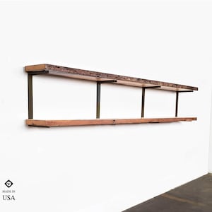 May include: Two wooden shelves with metal brackets mounted on a white wall. The shelves are made of reclaimed wood and have a rustic look. The metal brackets are a dark bronze color. The shelves are empty.