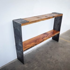 May include: A two-tiered shelf unit with a rustic wooden top and bottom shelf. The shelf is made of metal with a dark gray finish. The shelf is standing on a concrete floor.