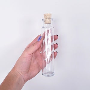 May include: A clear glass bottle with a cork stopper. The bottle is cylindrical and held in a hand against a white background. The cork is light brown. The bottle is empty.