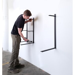 Puede incluir: Una persona instalando soportes de estanter&iacute;a de metal negro en una pared blanca con un taladro el&eacute;ctrico.