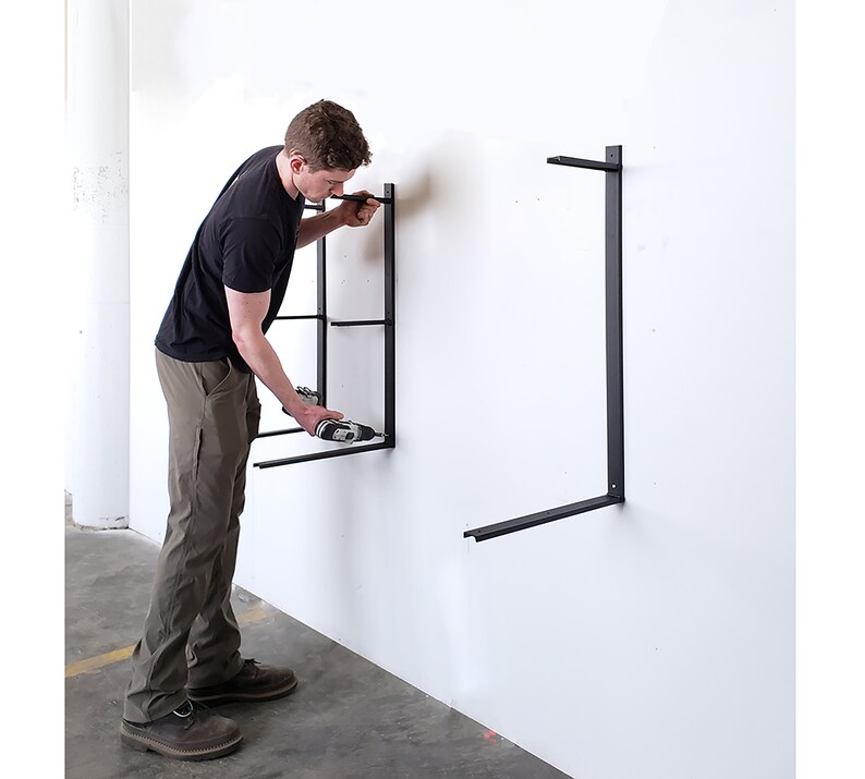 Puede incluir: Una persona instalando soportes de estanter&iacute;a de metal negro en una pared blanca con un taladro el&eacute;ctrico.