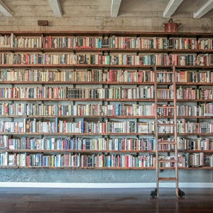 May include: A large wooden bookshelf filled with books of various sizes and colors. The bookshelf is mounted on a concrete wall and includes a wooden ladder for accessing the higher shelves. A decorative bucket sits on the top shelf.