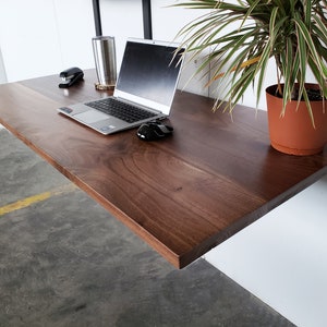 Puede incluir: Un escritorio flotante de madera marr&oacute;n oscuro con un port&aacute;til, un rat&oacute;n, una grapadora y una planta en maceta en la parte superior. El escritorio est&aacute; montado en una pared blanca.