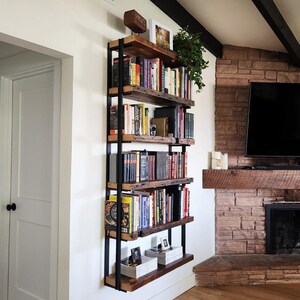 Pu&ograve; includere: Una libreria rustica a parete con cinque ripiani. I ripiani sono in legno di recupero e sostenuti da una struttura in metallo nero. Libri di varie dimensioni e colori riempiono i ripiani, insieme a oggetti decorativi e una piccola pianta.
