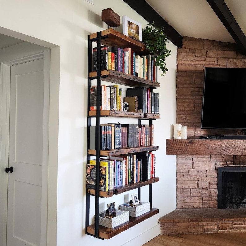 Pu&ograve; includere: Una libreria rustica a parete con cinque ripiani. I ripiani sono in legno di recupero e sostenuti da una struttura in metallo nero. Libri di varie dimensioni e colori riempiono i ripiani, insieme a oggetti decorativi e una piccola pianta.