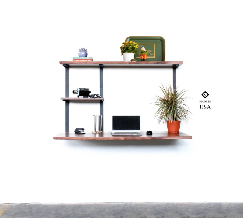 Puede incluir: Un escritorio flotante de nogal con soportes de metal negro y dos estantes. El escritorio tiene un port&aacute;til, una planta, una taza y una grapadora encima. Los estantes tienen una c&aacute;mara, gafas de sol y una peque&ntilde;a planta encima. El escritorio est&aacute; montado en una pared blanca. El texto "MADE IN USA" es visible en la imagen.
