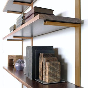 Pu&ograve; includere: Una moderna libreria a pi&ugrave; livelli con ripiani in legno scuro e supporti in metallo dorato. I ripiani espongono libri, blocchi di legno decorativi e una piccola pianta succulenta in vaso. Il design &egrave; minimalista ed elegante.