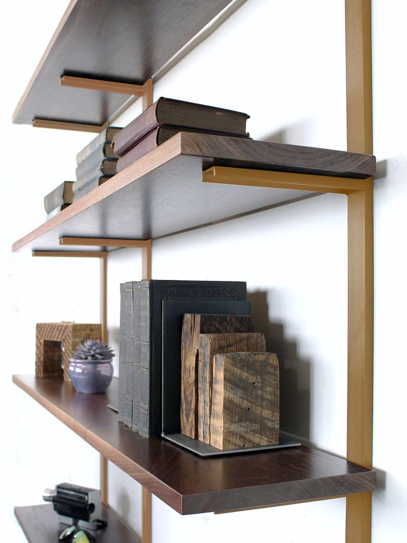 Pu&ograve; includere: Una moderna libreria a pi&ugrave; livelli con ripiani in legno scuro e supporti in metallo dorato. I ripiani espongono libri, blocchi di legno decorativi e una piccola pianta succulenta in vaso. Il design &egrave; minimalista ed elegante.