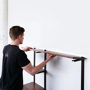 Puede incluir: Una persona instalando una estanter&iacute;a de madera en una unidad de estanter&iacute;a de pared de metal negro. La estanter&iacute;a est&aacute; hecha de madera oscura y se coloca cuidadosamente sobre los soportes.