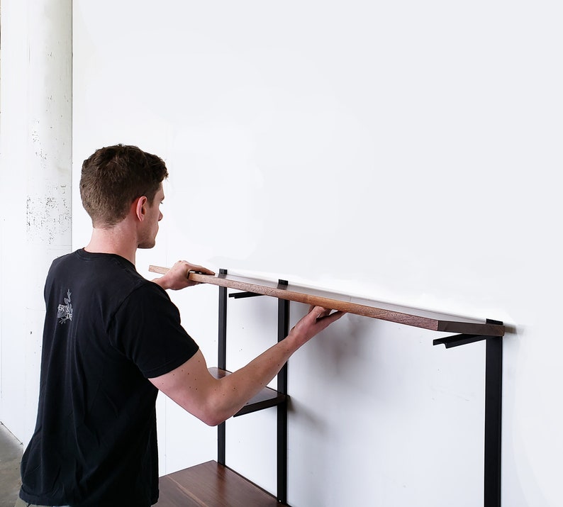 Puede incluir: Una persona instalando una estanter&iacute;a de madera en una unidad de estanter&iacute;a de pared de metal negro. La estanter&iacute;a est&aacute; hecha de madera oscura y se coloca cuidadosamente sobre los soportes.