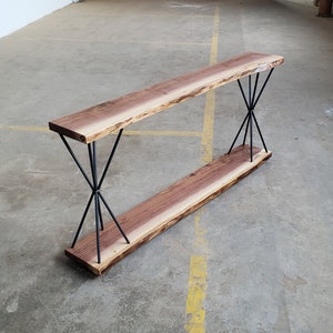 May include: A modern console table with a natural wood top and bottom shelf. The wood has a live edge and a rich brown color. The table is supported by black metal legs that form an X shape, creating a unique and stylish design.