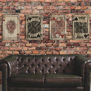 May include: Four vintage-style playing cards, featuring the Ace, Jack, Queen, and King of Spades, are displayed on a brick wall. The cards are printed on wood and have a distressed, aged look. The cards are arranged in a row, with the Ace on the left and the King on the right. A brown leather sofa is in the foreground.
