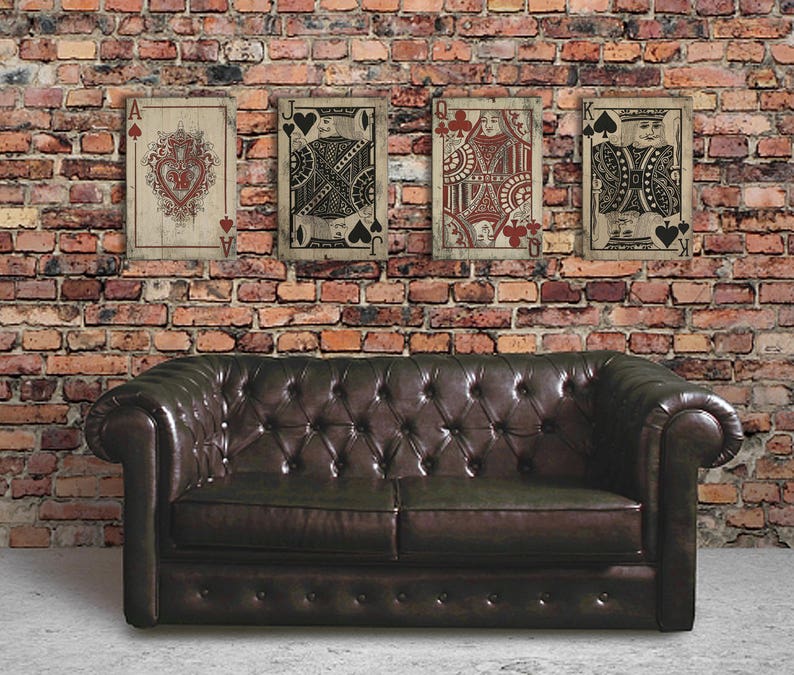 May include: Four vintage-style playing cards, featuring the Ace, Jack, Queen, and King of Spades, are displayed on a brick wall. The cards are printed on wood and have a distressed, aged look. The cards are arranged in a row, with the Ace on the left and the King on the right. A brown leather sofa is in the foreground.