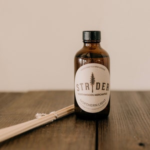 May include: A brown glass bottle with a label that reads "STRIDER NORTHWOODS MERCANTILE NORTHERN LIGHT" and a small pine tree graphic. The bottle is sitting on a wooden surface with a set of wooden diffuser sticks.