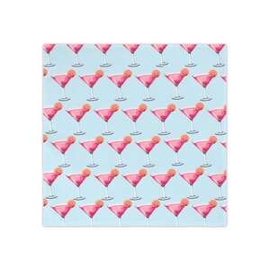Op de afbeelding: Een lichtblauwe stof met een herhalend patroon van roze cocktails in glazen op een voet. Elk glas is gegarneerd met een schijfje sinaasappel op de rand. Het ontwerp is in een raster over het gehele oppervlak van het materiaal geplaatst.