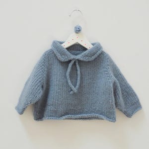 Può includere: Un maglione in maglia a maniche lunghe color blu polvere caratterizzato da un ampio colletto con chiusura a laccetti coordinata sullo scollo. Il maglione è appeso a una gruccia bianca decorata con piccoli puntini rossi e un pon-pon blu.
