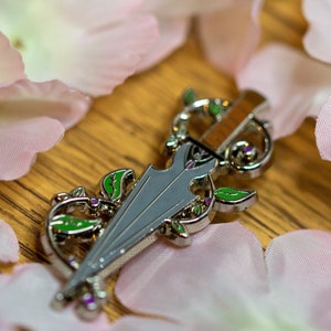 May include: A silver and green enamel pin shaped like a dagger with floral details. The pin is surrounded by pink flower petals.