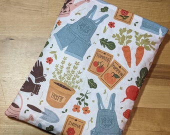 May include: A white fabric book sleeve featuring a garden-themed pattern. The design includes light blue overalls, seed packets labeled "Tomato," "Pumpkin," and "Zinnia," carrots, and a pot of thyme. Other elements include a watering can, gardening gloves, and a trowel.