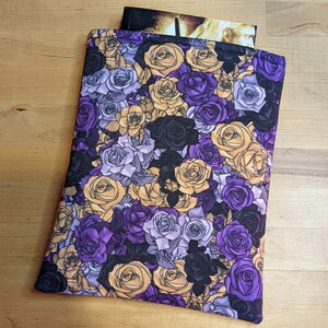 Peut inclure: Une housse de livre florale violette et dorée avec un motif de roses. La housse est en tissu et possède une fermeture à glissière.