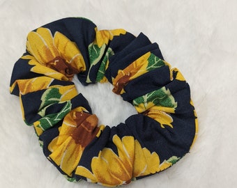 May include: A fabric scrunchie with a dark blue background and a pattern of yellow sunflowers with green leaves. The scrunchie is circular and gathered, designed to hold hair.
