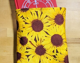 May include: A red book with a white clock face design on the cover is inside a book sleeve with a sunflower pattern. The sleeve is made of fabric and has a brown background with yellow sunflowers.