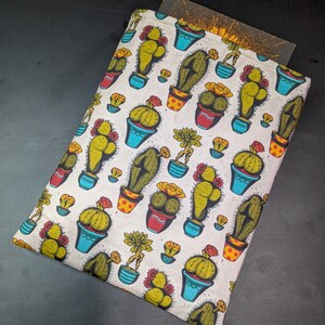 May include: A white fabric book sleeve featuring a repeating pattern of colourful cacti in pots. The cacti are in various shapes and sizes, with some having flowers.