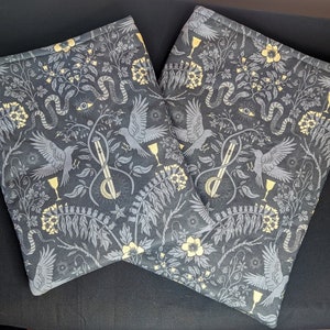May include: Two black fabric book covers with a gold and grey floral pattern. The pattern includes birds, snakes, and chalices.