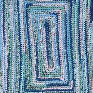 May include: A rectangular rug with a braided design. The rug features a pattern of concentric rectangles in shades of blue, green, and pink. The rug is made of fabric strips and has a handmade appearance.