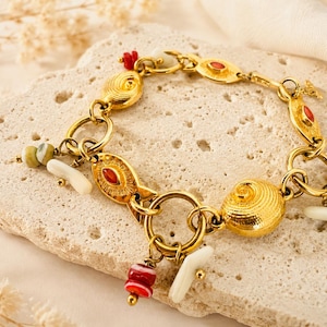 Puede incluir: Una pulsera de dijes en tono dorado con una serie de eslabones circulares interconectados. La pulsera está adornada con varios dijes, incluyendo formas de peces texturizados, una concha en espiral y pequeñas cuentas en tonos rojo, blanco y verde.