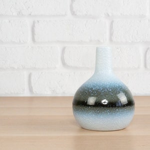 May include: A decorative ceramic vase with a rounded body and a narrow neck. The vase features a gradient of colors, transitioning from white at the base to blue and black near the middle, and back to white at the top. The vase is on a wooden surface.
