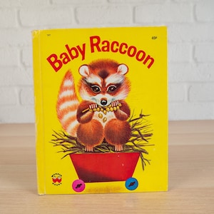 May include: A vintage children's book titled "Baby Raccoon" with a yellow cover. The cover features a cartoon raccoon holding food, set in a red wagon. The book is from Wonder Books.