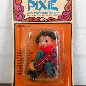 May include: A vintage Pixie air freshener in original orange blister packaging. The product features a small doll with a red scarf, plaid shirt, and dark pants. The card displays the text Pixie Air Refresher for use in home or auto.