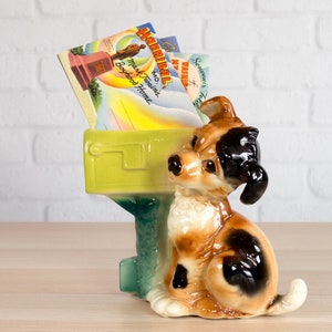 May include: A ceramic dog-shaped mailbox in shades of brown, black, and white, with a green mailbox section. The mailbox is filled with vintage postcards. The dog is sitting upright, with its head turned to the side.