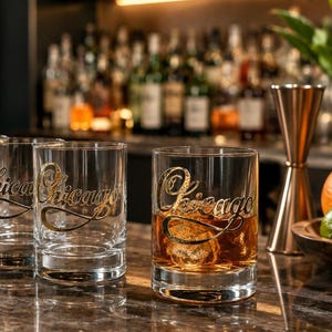 May include: Four clear glass tumblers arranged in a row, each featuring the word Chicago etched in a cursive script. The rightmost glass contains an amber-colored liquid with ice cubes, while the others are empty. The glasses have thick bases.