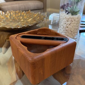May include: A square wooden box with rounded corners and a natural wood finish. The box has a rectangular opening on top with a black insert. The box is sitting on a glass table.