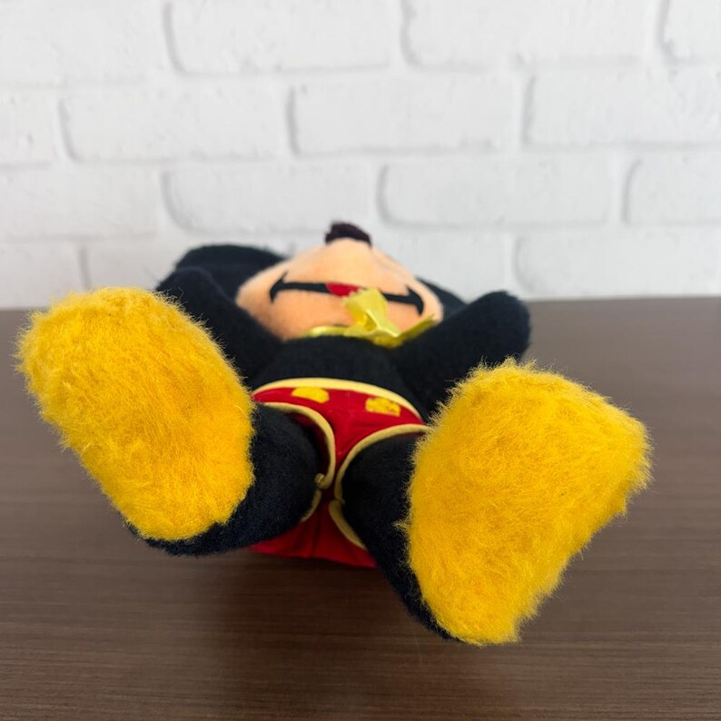 May include: A Mickey Mouse soft toy lying on its back. The toy has black fur, large yellow feet, and red shorts with yellow details. Mickey's face is visible, with a red mouth and a yellow bow.
