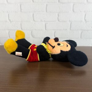 May include: A plush Mickey Mouse toy, lying on its back. The toy features black fur, yellow shoes and gloves, red shorts, and a yellow collar. The face is tan with black eyes and a brown nose.