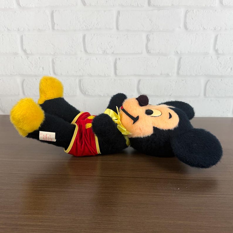May include: A plush Mickey Mouse toy, lying on its back. The toy features black fur, yellow shoes and gloves, red shorts, and a yellow collar. The face is tan with black eyes and a brown nose.