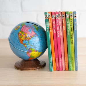 May include: A blue globe bank with a colorful world map sits next to a collection of vintage children's books. The books have colorful spines and titles like "My Pets" and "Monkey Play". The globe is approximately 6 inches in diameter.