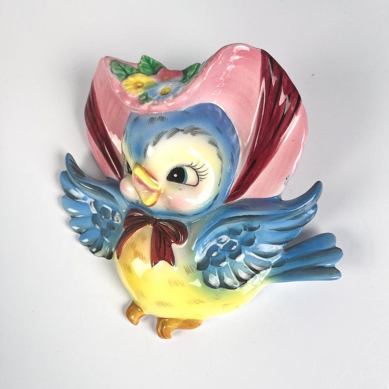 May include: A ceramic wall plaque shaped like a blue bird with a yellow belly and a dark red bow tie. The bird wears a large pink bonnet adorned with small yellow, blue, and pink flowers. The wings are spread wide, and the bird has large eyes.