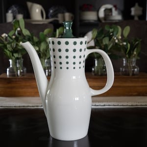 May include: A white ceramic coffee pot with a long spout and a handle. The top of the pot and the lid are dark green. The upper portion of the pot has a pattern of evenly spaced dark green dots. The pot is on a dark wooden table.