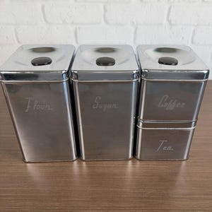 May include: A set of four rectangular chrome canisters with black circular knobs on the lids. The canisters are labeled with etched text reading Flour, Sugar, Coffee, and Tea. The containers have a metallic finish and a vertical orientation.